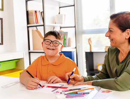 A child and carer colouring on paper together. The child is smiling to camera, the carer smiling toward the child